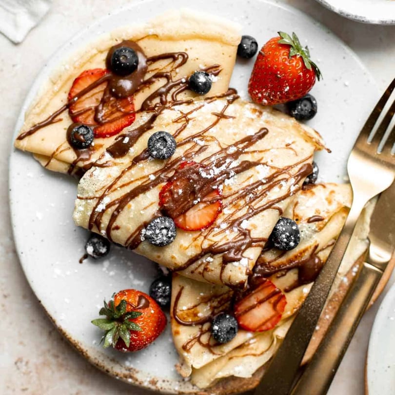 Folded crepes drizzled with chocolate, powdered sugar, fresh strawberries, and blueberries on a plate.