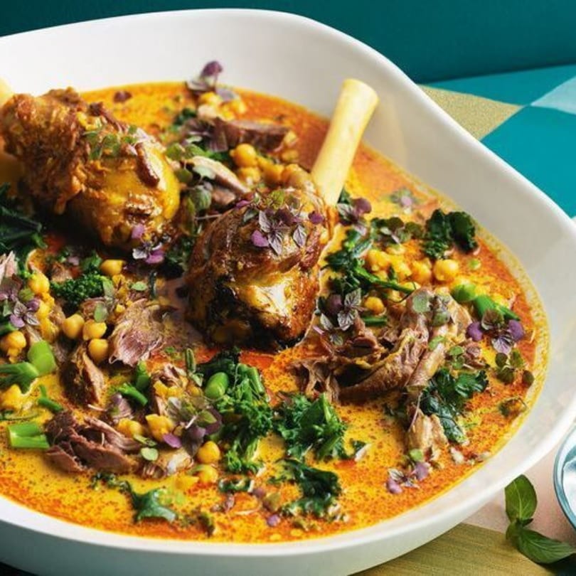 Mediterranean lamb shank stew with chickpeas, greens, and purple herbs in a white bowl.