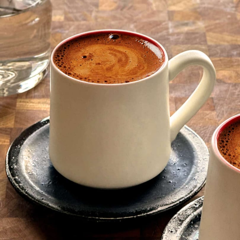 A white mug of rich, foamy Turkish coffee sits on a dark saucer next to a glass of water.