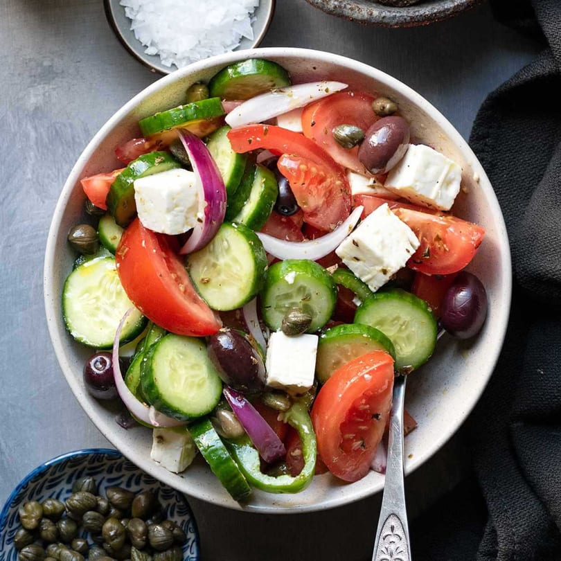 A vibrant Mediterranean salad with cucumbers, tomatoes, feta, olives, and red onion in a white bowl.